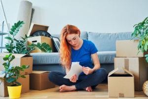 woman sitting on floor reading pages in living room around couch and moving boxes